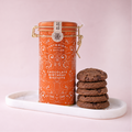 An orange metal canister of Cartwright Butler Chocolate Birthday Biscuits sits on a white marble tray next to a stack of chocolate chip cookies against a soft pink background.
