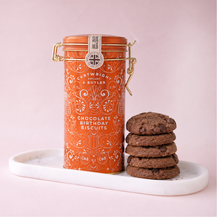 An orange metal canister of Cartwright Butler Chocolate Birthday Biscuits sits on a white marble tray next to a stack of chocolate chip cookies against a soft pink background.
