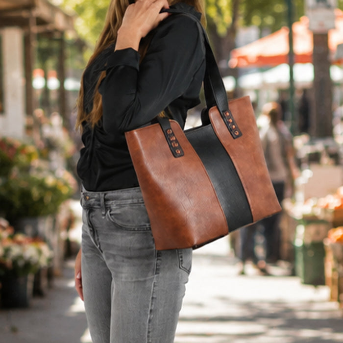 A woman wearing a black long-sleeve top and gray jeans carries a large brown faux-leather tote bag with a wide black center stripe and black shoulder straps in an outdoor street market setting. Concealed carry Wrenly Tote purse with Aztec pattern and magnetic firearm pocket