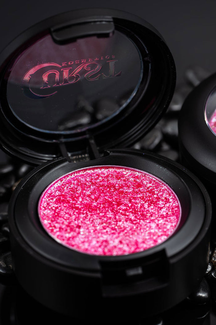 Compact makeup case with pink glittery eyeshadow and visible curst brand logo on a dark background