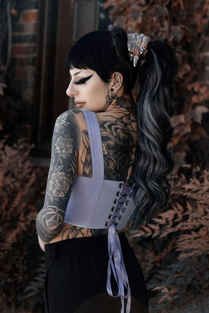 Person wearing a light purple corset with intricate designs, standing against a textured wall.