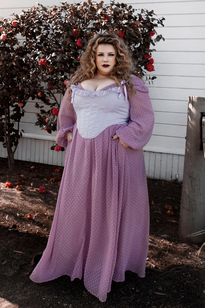 Woman in a purple dress and corset standing in front of a white building with a bush bearing red fruits.