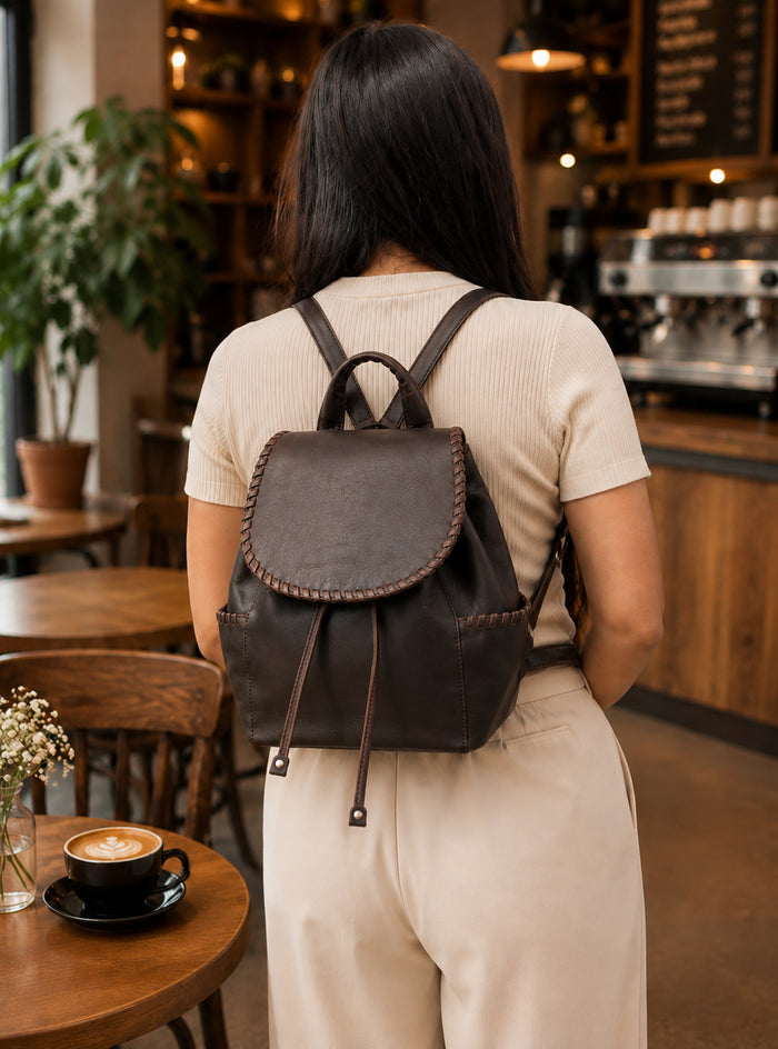 A woman viewed from behind wearing a dark brown leather backpack with drawstring straps, standing in a cozy, warmly lit coffee shop with wooden tables and shelves in the background.