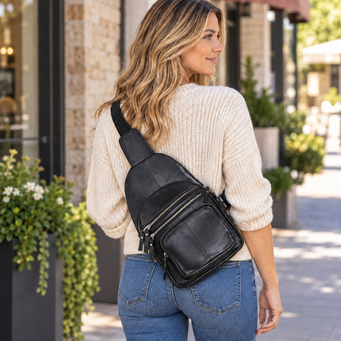 A woman wearing a cream knit sweater and blue jeans is shown from the back, carrying a black leather crossbody bag with multiple zippered pockets on a sunny outdoor sidewalk. Concealed Carry Weston Lambskin Sling by DS Conceal - Universal Holster Backpack - Leather Pistol CCW Bag - Unisex Pistol Bag - Ambidextrous Holster Bags Lady Conceal Firearm Bags - Self Defense Bag for Gun