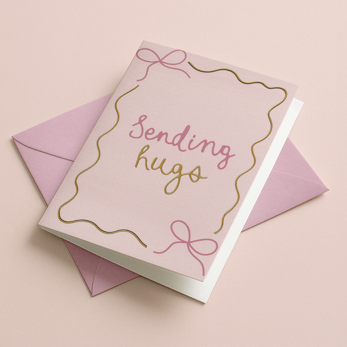 A pink greeting card with the words 'Sending hugs' in pink and gold script, decorated with pink bow outlines, shown with a matching pink envelope on a soft pink background.