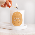 A white candle in a white glass holder sits on a white tray, with a hand lighting a match above it. The front of the candle features a tan oval graphic with the text 'SMELLS LIKE YOU'VE GOT THIS' and small star accents.