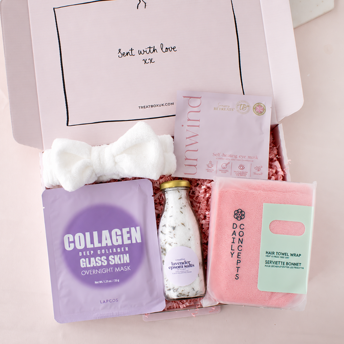 A curated skincare gift box from Treat With Love, featuring a white plush bow, a lavender collagen mask packet, a bottle of lavender epsom salts, a pink hand towel wrap, and a pink bath towel wrap, arranged in pink shredded paper inside an open pink box.
